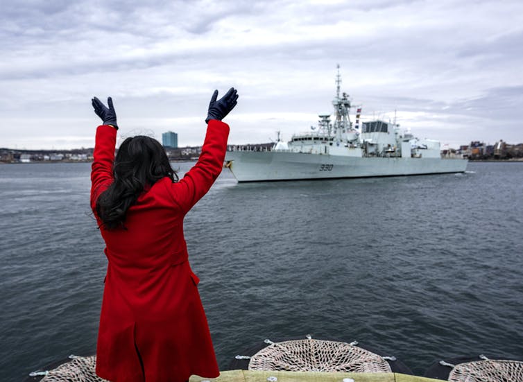 Une femme en manteau rouge aux cheveux noirs est vue de dos alors qu'elle salue un navire de patrouille dans un port.