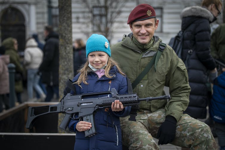 Une fille dans un bonnet bleu vif tient un fusil militaire alors qu'un soldat en uniforme dans un béret rouge foncé sourit assis à côté d'elle.