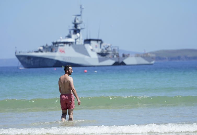 Un homme en maillot de bain rouge se tient dans les vagues avec un grand navire de patrouille en arrière-plan.